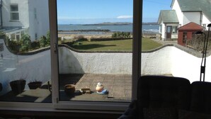 A view from a room with a sliding glass door to a patio area. The patio has a small table with a bowl and a plant. There are potted plants on the patio and a chair in the foreground. - Bright modern holiday cottage steps from the beach and in the heart of village (Rhosneigr)