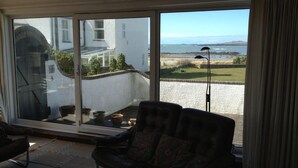 A room with a large sliding glass door leading to a balcony with a view of the sea and a white building. There is a black leather sofa with patterned cushions on the balcony. - Bright modern holiday cottage steps from the beach and in the heart of village (Rhosneigr)