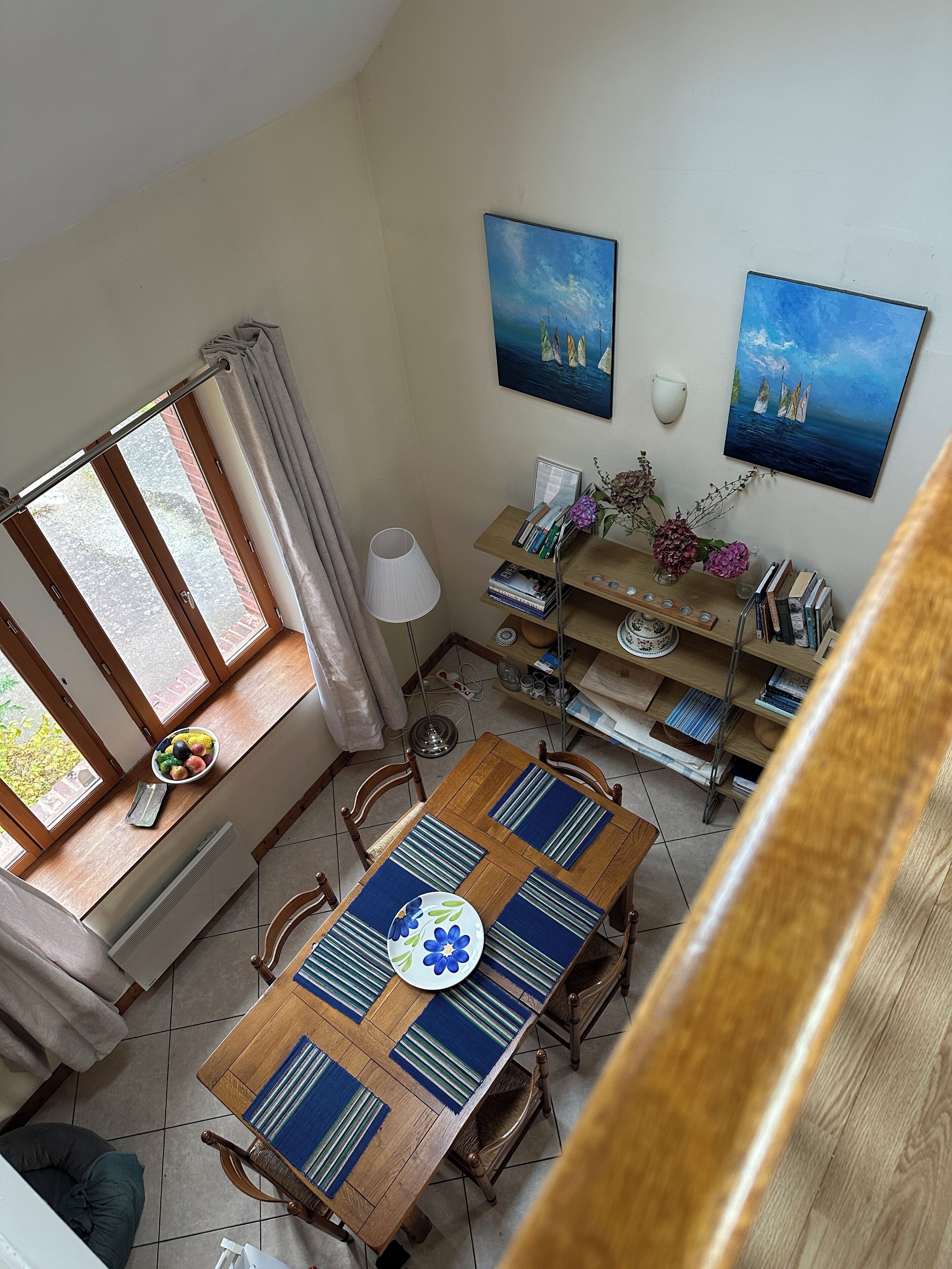 A dining area with a wooden table, chairs, a window with curtains, a bookshelf, and two paintings on the wall.