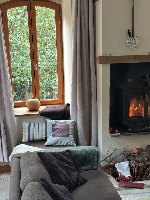 A cozy room with a sofa, a window with curtains, a fireplace, and a view of greenery outside.
