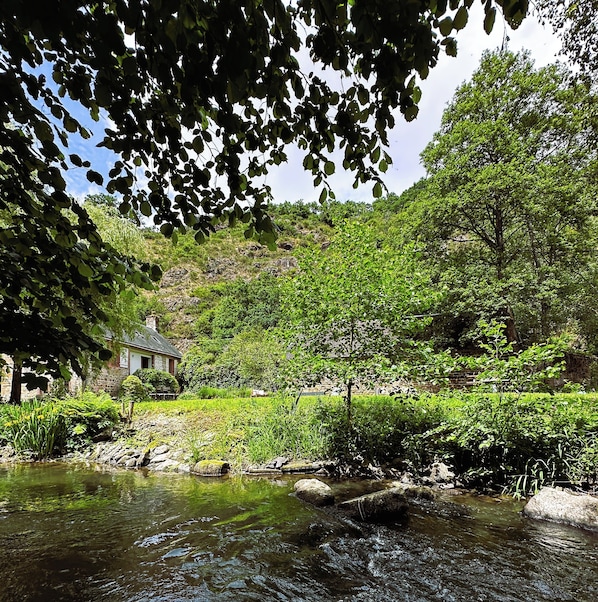 A stone building with a sloped roof, surrounded by lush greenery and a stream.