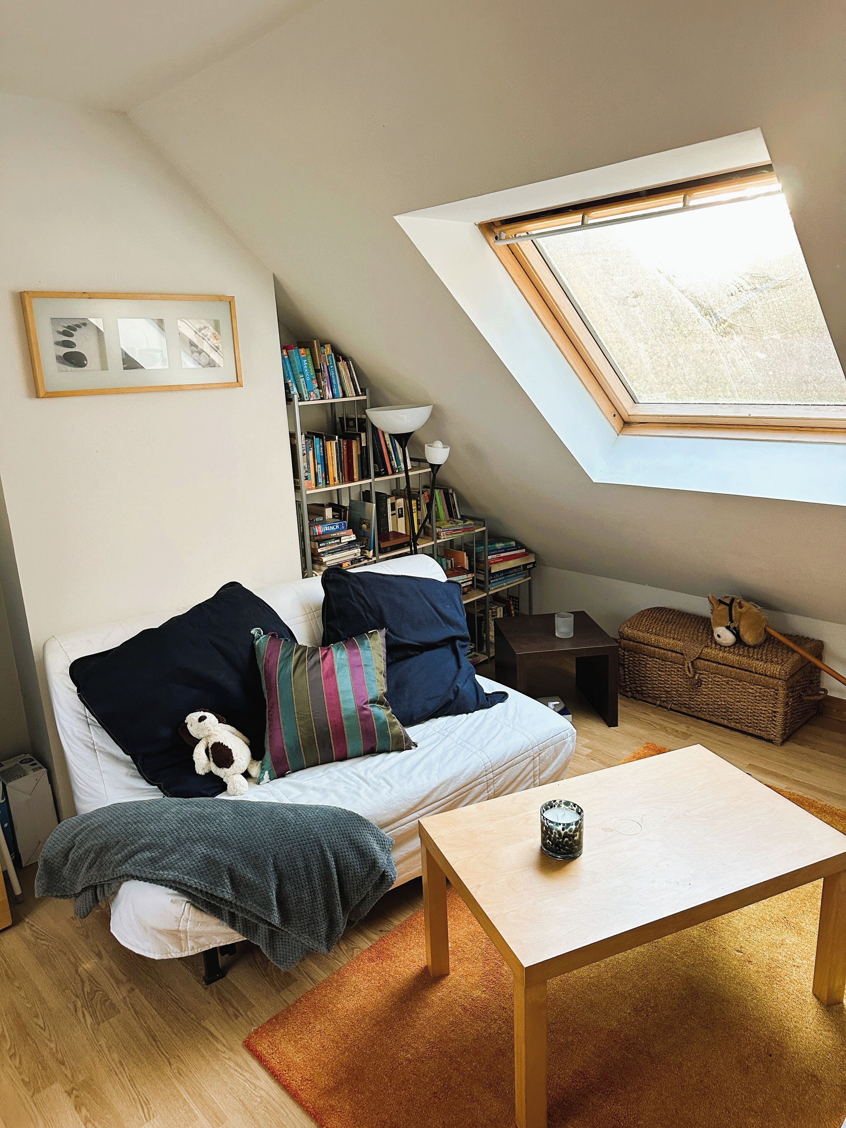 Een knus slaapkamerloft met een schuine kap, dakraam, een boekenkast, een kleintje tafeltje en een comfortabele bank.