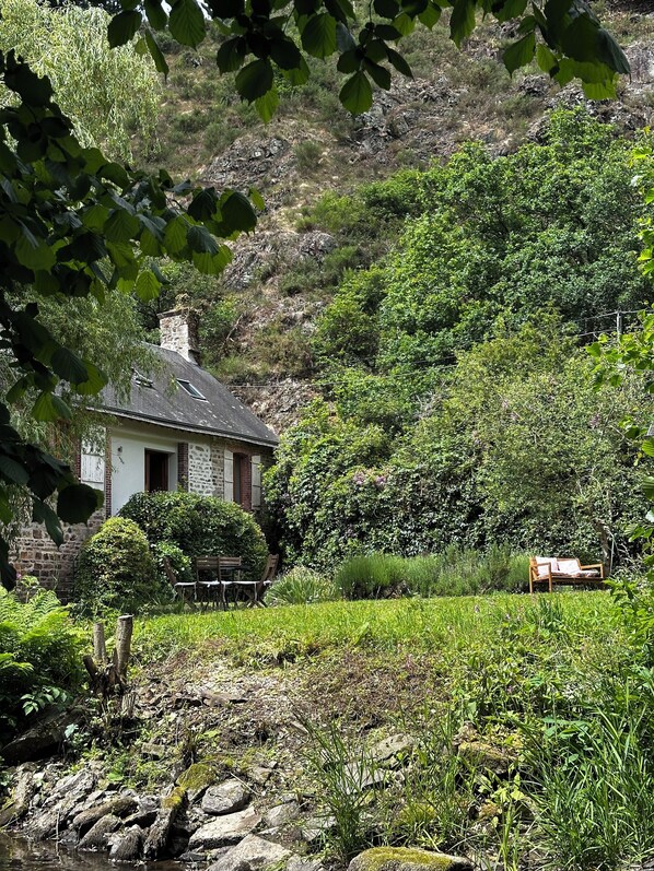 A stone house with a chimney, surrounded by lush greenery and a small pond.