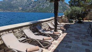 A seaside terrace with lounge chairs and a stone wall overlooking the sea and mountains.