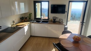 A modern kitchen with a large island, a dining area with a table and chairs, a flat-screen TV mounted above the island, and a view of the outdoors through the open doors. - South facing Villa with Panoramic Sea View, POOL, AC, Quiet  (Grasse)