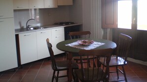 Une cuisine compacte avec des meubles blancs, un évier et une cuisinière. On y trouve une table à manger avec des chaises et une fenêtre avec des stores.