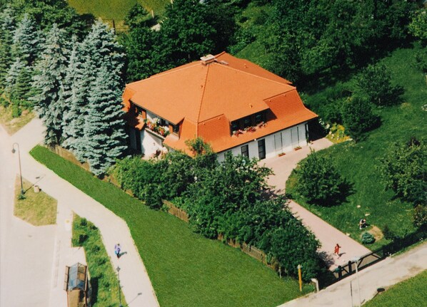 An aerial view of a house with a red roof, surrounded by greenery and a paved driveway.