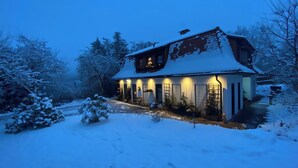 Una casa cubierta de nieve con un porche visible y una ventana iluminada, rodeada de árboles nevados.