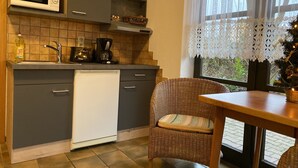 Cocina compacta equipada con microondas, cafetera y lavavajillas. Hay una silla de mimbre y una pequeña mesa de comedor cerca de una ventana con cortinas de encaje.