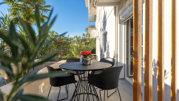 An outdoor patio with a round table and four chairs, surrounded by palm trees and a building with large windows. - Close to the beach, quiet location, family-friendly (Funtana)