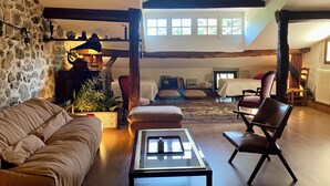 Un salon chaleureux avec un mur en pierre, des poutres en bois, un canapé, une table basse et une vue sur la chambre à coucher par une porte ouverte.