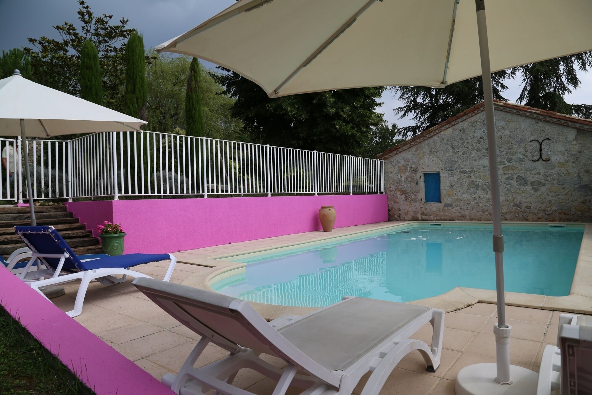 A swimming pool with a white fence, a pink wall, and a stone building with a blue door in the background.