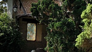 Un bâtiment rustique avec un toit en tuiles, un mur en pierre et une fenêtre ornée d’un motif en losanges. On y trouve des plantes en pot et un banc en pierre devant.