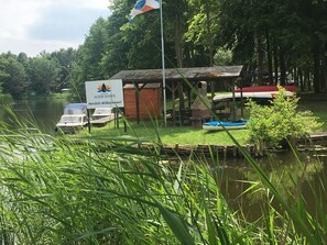 A riverside cabin with a sign reading "MARINA ALTER HAEN Herzlich Wilkommen!" and a flag. Boats are docked nearby. - Stay overnight close to nature - the camping barrel (Mildenberg)
