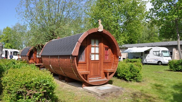 Ein baubares, fassförmiges Gebäude mit Tür und Fenstern, umgeben von Grünflächen und weiteren Wohnmobilen.