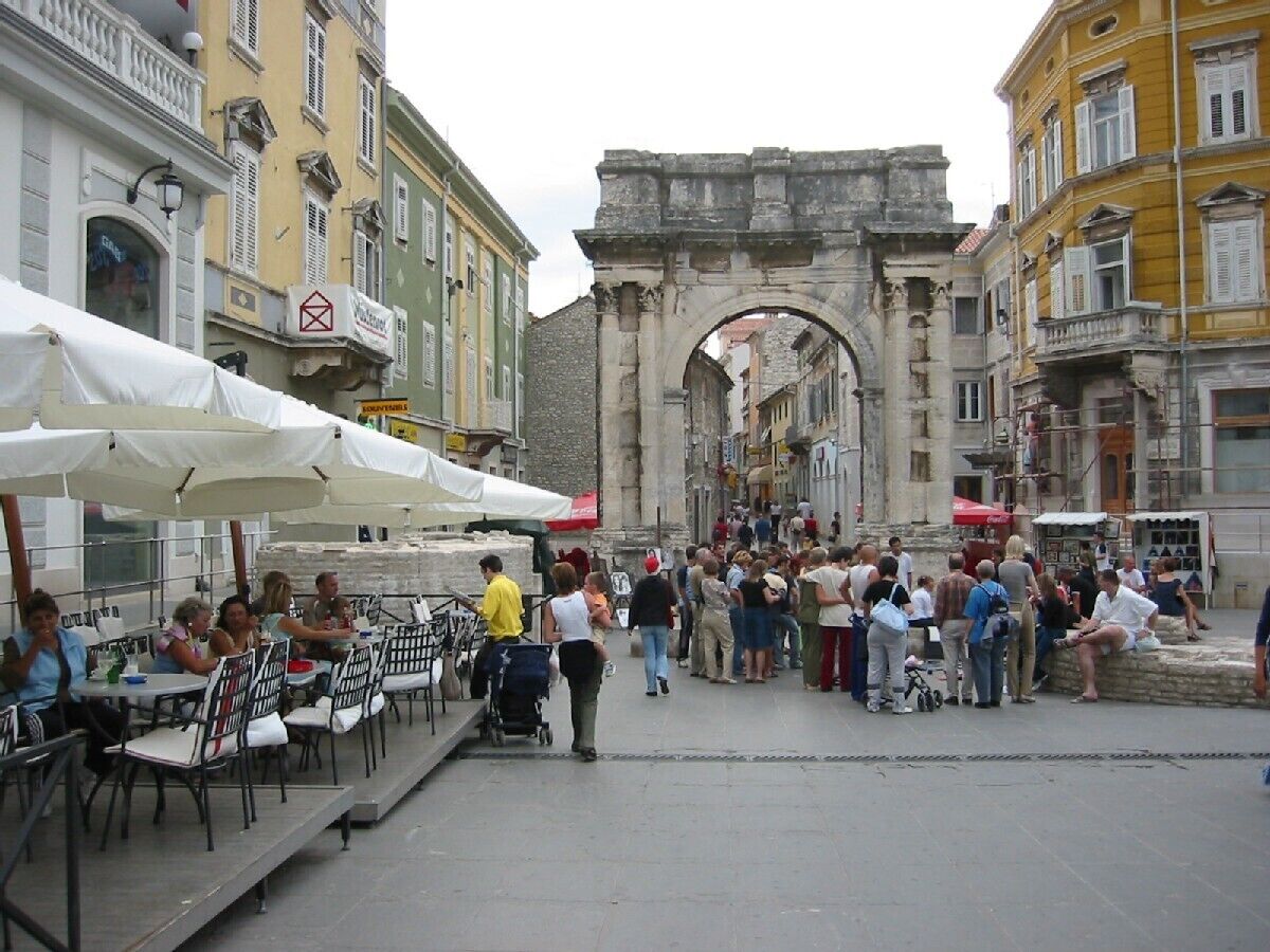 An outdoor café with tables and umbrellas, a stone archway in the background, and people sitting and walking.