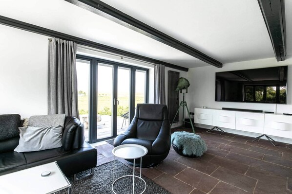 Un salón moderno con un sofá de cuero negro, un sillón negro, una mesita redonda pequeña, una lámpara de pie y un televisor montado en la pared.