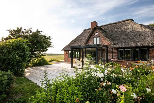 A thatched-roof house with a glass conservatory, surrounded by lush greenery and a wooden deck.