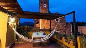 Una hamaca bajo una pérgola de madera, un edificio de piedra con una torre de reloj y un telón de fondo montañoso.