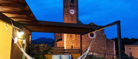 A hammock under a wooden pergola, a stone building with a clock tower, and a mountainous backdrop.