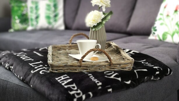 A modern living room with a grey sofa, decorative pillows, a woven tray with a vase of flowers, and a cushion with text.