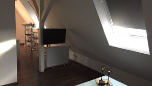 A modern loft-style room with a sloped ceiling, a flat-screen TV mounted on a white wall, a dining area with a table and chairs, and a small table with a remote control and two candles.