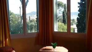 A room with a window overlooking a scenic landscape, a small table with a clear glass vase, and a bed with a red pillow. - House / Villa - SANARY SUR MERHouse with character (Sanary-Sur-Mer)