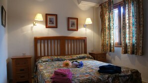 Ein Schlafzimmer mit einem hölzernen Kopfende, zwei Nachttischen, einem Bett mit floralem Überwurf, zwei Wandlampen und einem Fenster mit gemusterten Vorhängen.