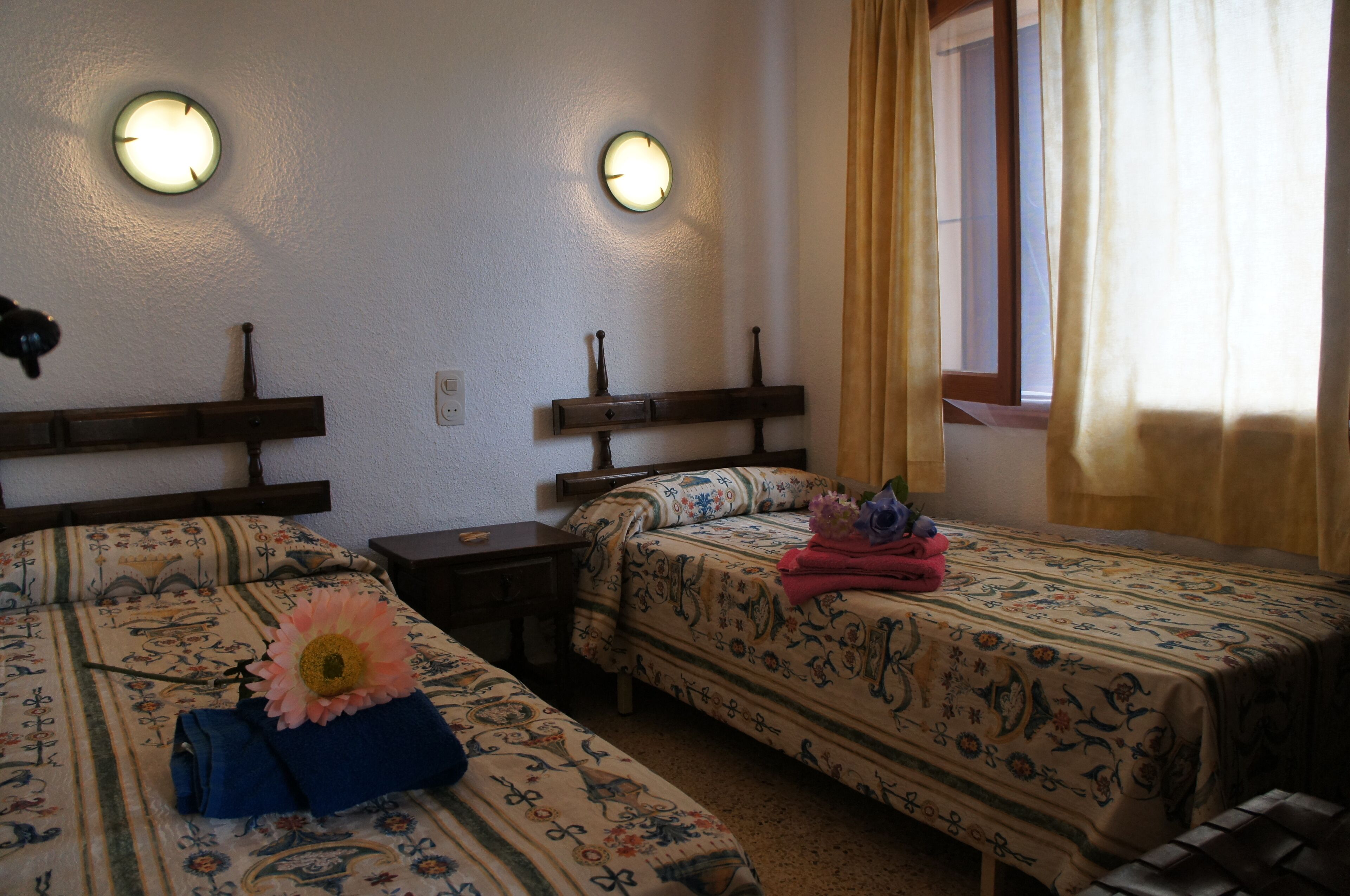 A hotel room with two beds, each with floral-patterned bedding, a nightstand, and a wall-mounted clock.