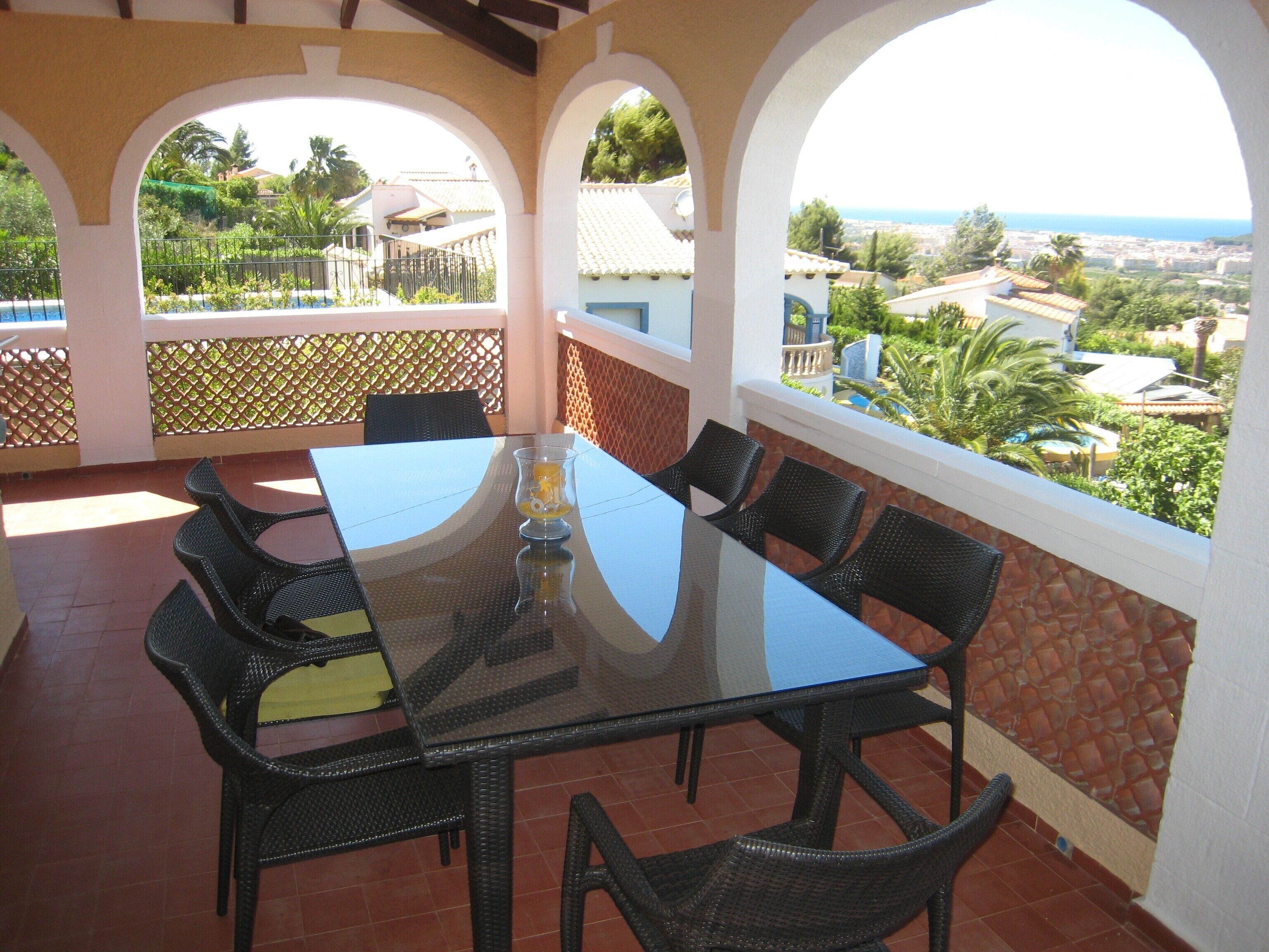 A glass-top dining table with wicker chairs, set on a patio with a view of a coastal town and greenery.