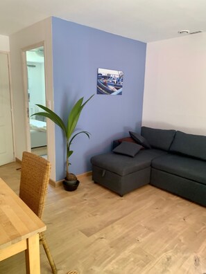 A living room with a grey sofa, a wooden dining table, a wicker chair, a potted plant, and a wall-mounted picture.