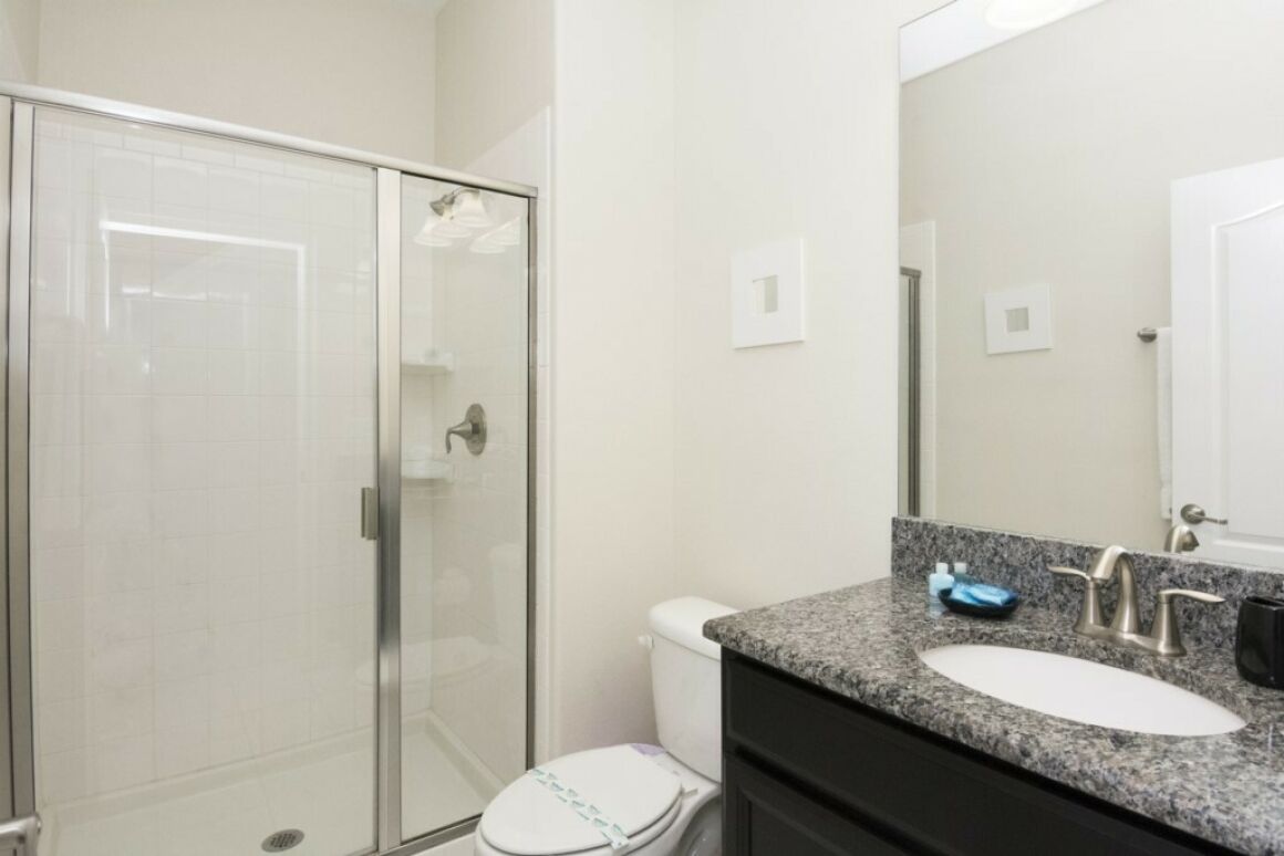 Un baño con cabina de ducha de vidrio, inodoro blanco, lavabo con encimera de granito y espejo.