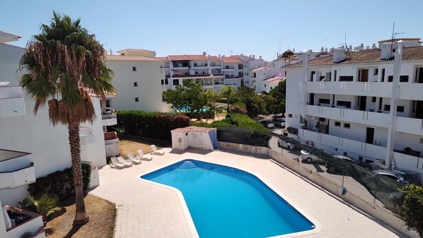 A swimming pool surrounded by white buildings with balconies, palm trees, and a clear sky.
