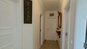 A hallway with wooden flooring, white walls, and doors. There is a decorative mirror on the right wall and a framed artwork above one of the doors.