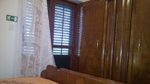 Un dormitorio con cama, armario de madera, cortinas de encaje y ventana con persianas.
