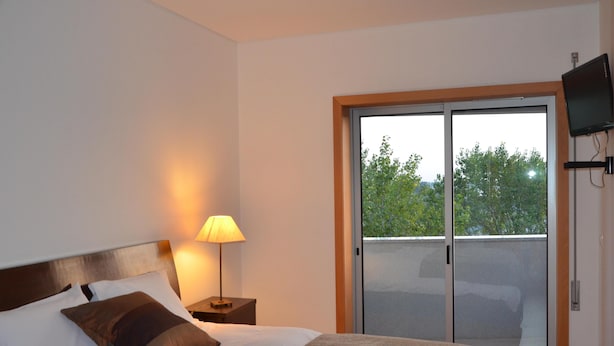 Une chambre d’hôtel comprenant un lit, une table de chevet avec une lampe, une porte-fenêtre coulissante donnant sur un balcon et un téléviseur fixé au mur.