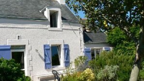 Ein zweistöckiges Haus mit einer kleinen Veranda, einem Schornstein und einem Garten mit verschiedenen Pflanzen und einer Bank.