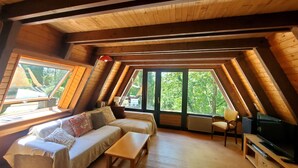 Un acogedor dormitorio en el ático con sofá, mesa y televisor. Amplias ventanas ofrecen vistas al verdor exterior.