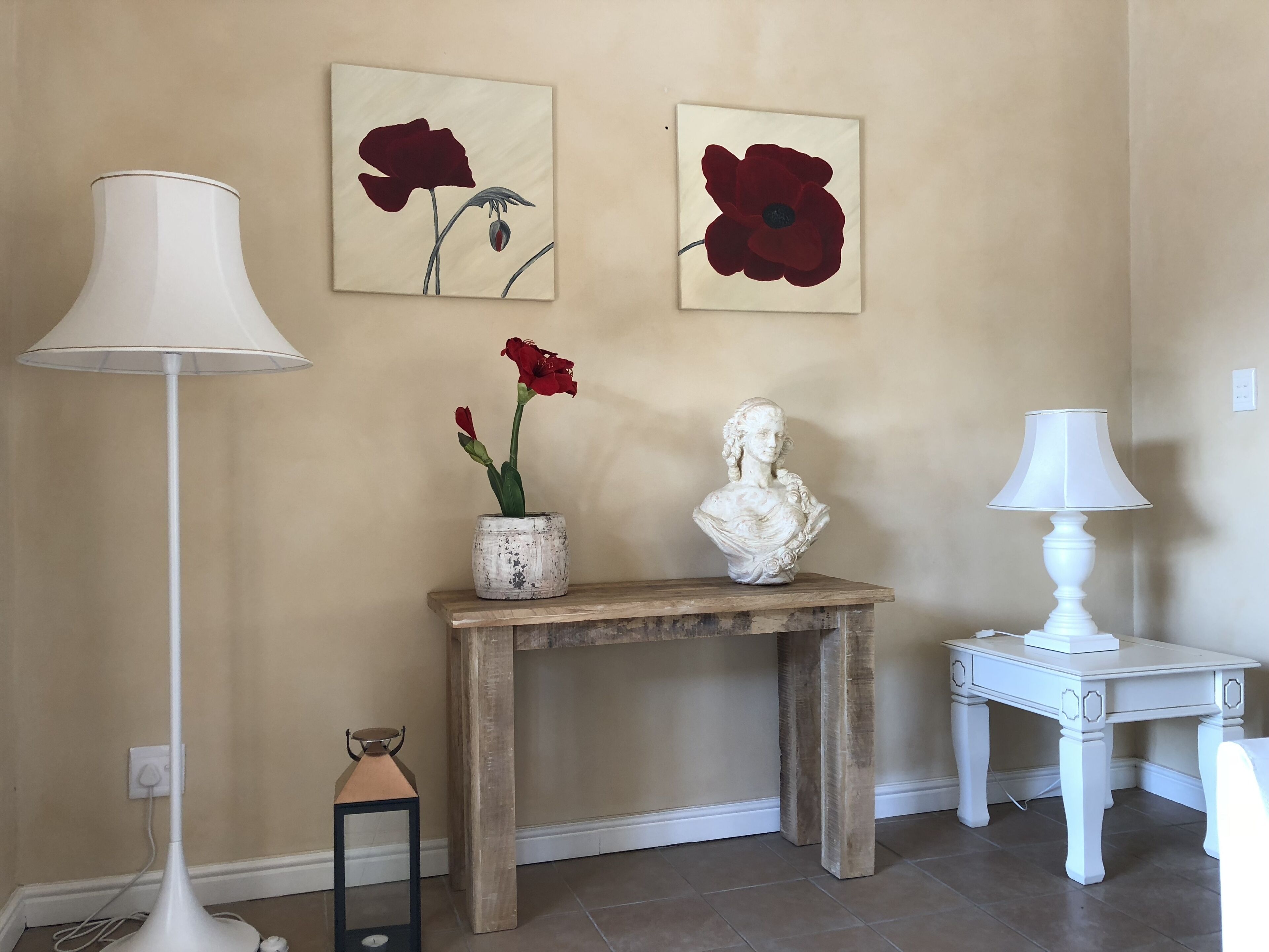Una habitación con una consola de madera, una escultura de busto, un jarrón con flores rojas, una lámpara de pie y dos cuadros colgados en la pared.