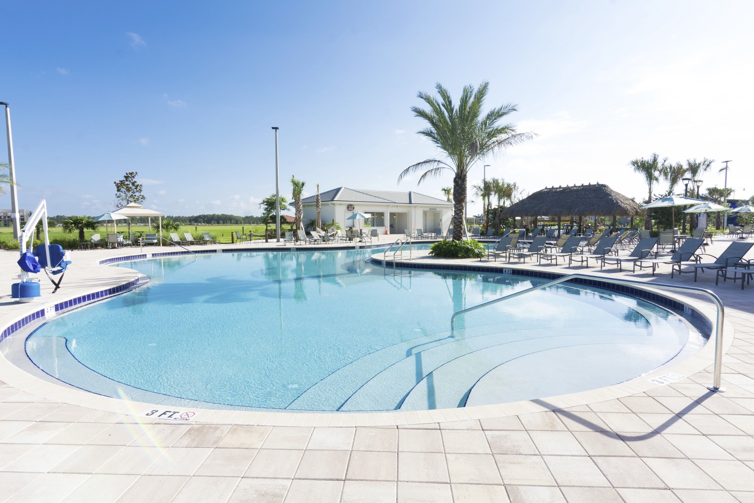 A swimming pool with lounge chairs and umbrellas, surrounded by palm trees and a building in the background.