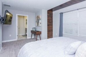 A bedroom with a large bed, a wooden accent wall, a desk with a chair, a mirror, and a window with blinds.