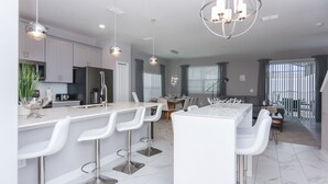 A modern kitchen with white bar stools, stainless steel appliances, and pendant lighting.
