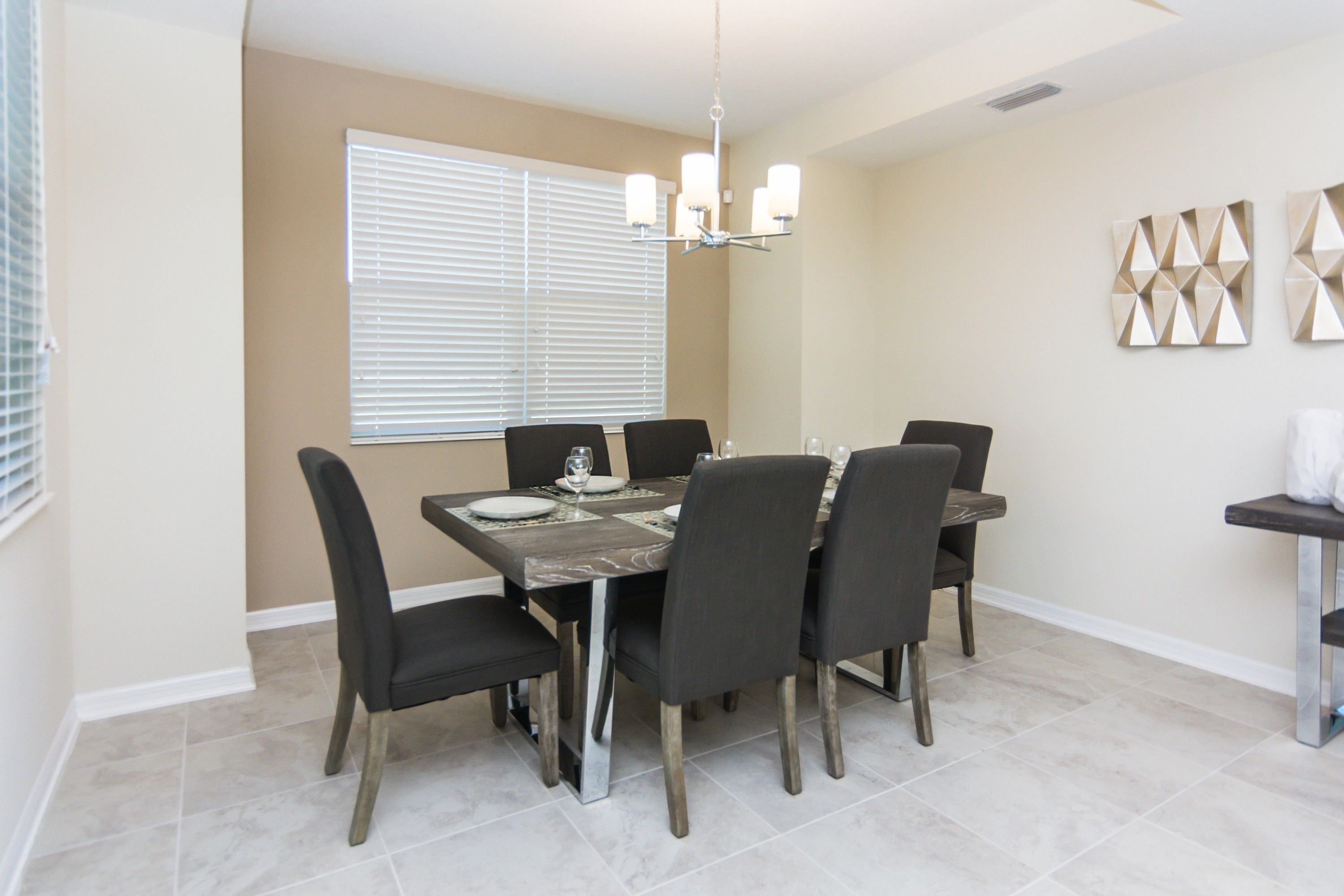 A dining area with a rectangular table, six chairs, a chandelier, and a window with blinds.