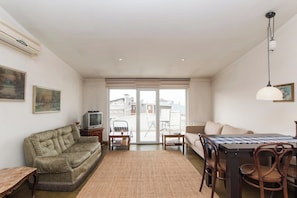 Uma sala de estar com sofá, televisão, mesa de jantar com cadeiras e uma janela grande com vista para edifícios.