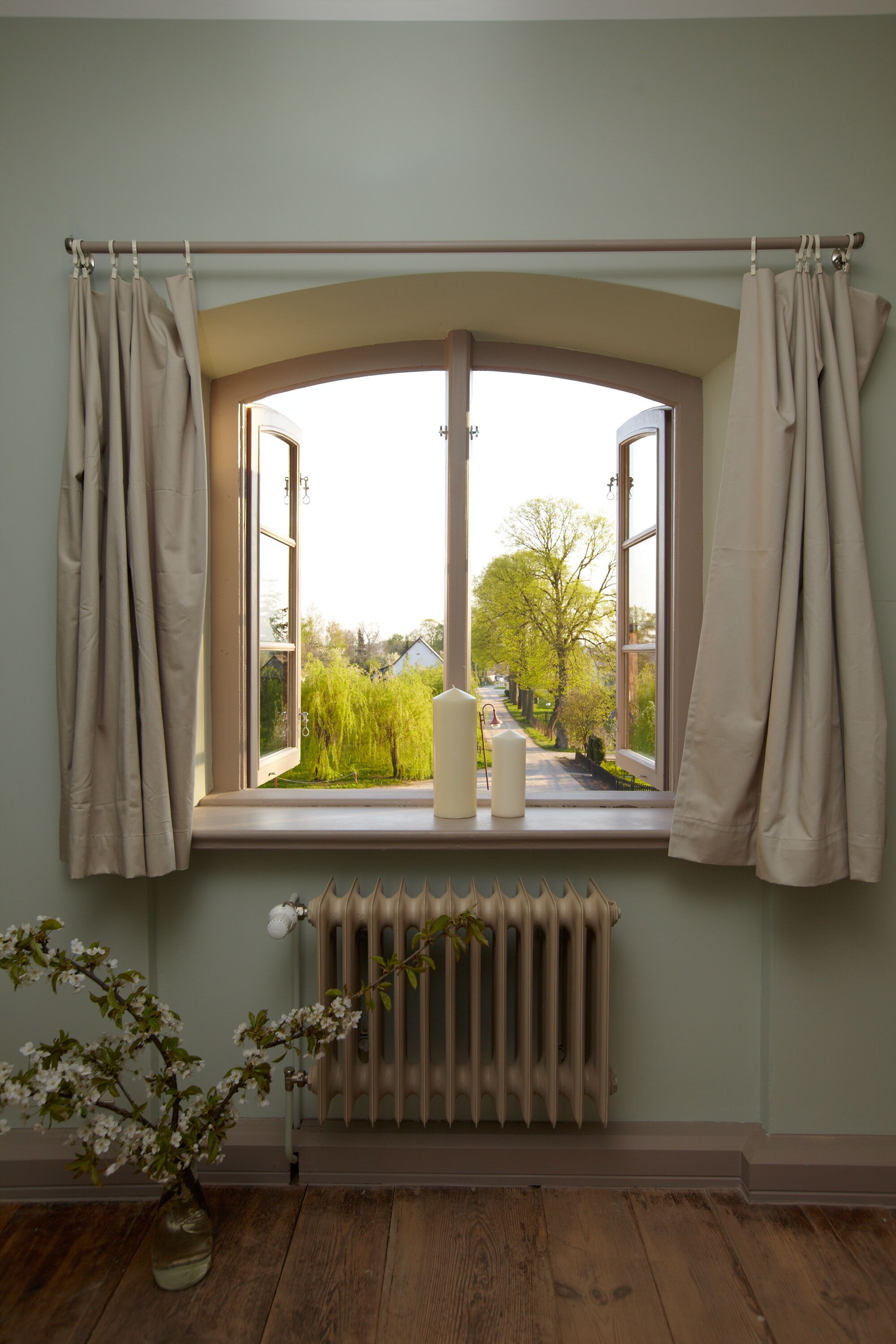 Ein Zimmer mit einem großen Fenster, einem Heizkörper darunter und einer Vase mit Blumen auf dem Boden.