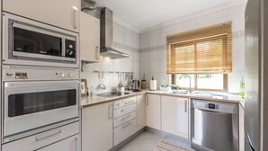 Una cocina moderna con electrodomésticos de acero inoxidable, armarios blancos y un ventanal con persianas.