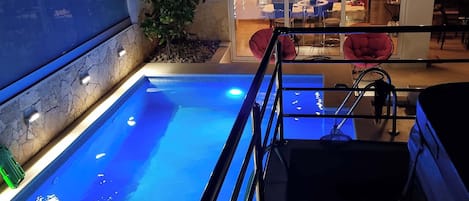 A swimming pool with a metal ladder, surrounded by a stone wall and a small garden area.