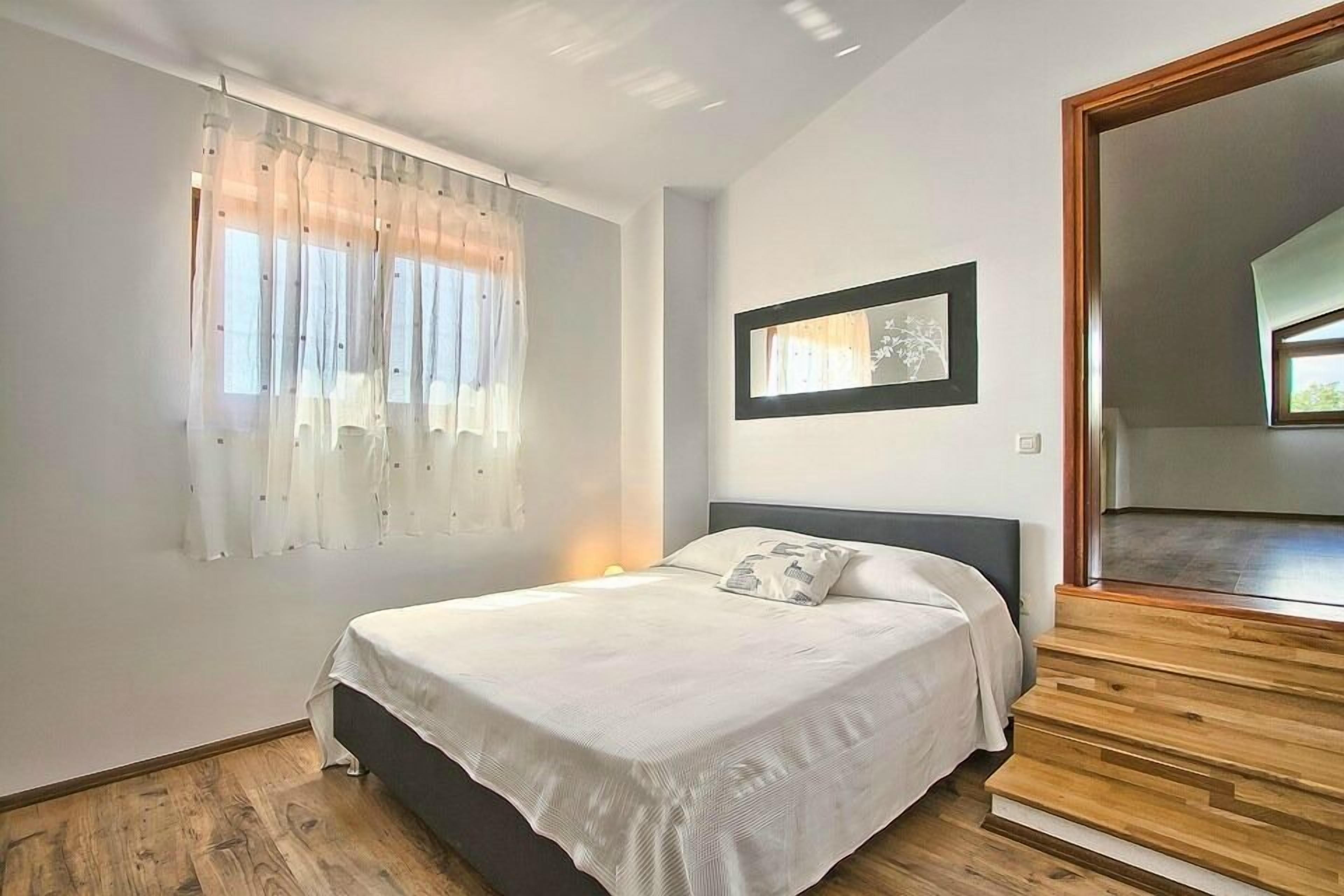 Ein Schlafzimmer mit einem Bett, einem Fenster mit leichtem Stoffvorhang, einem gerahmten Bild an der Wand und einer Holztreppe.