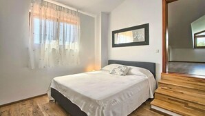 Een slaapkamer met een bed, een raam met dunne gordijnen, een lijst met een afbeelding aan de wand en een houten trap.