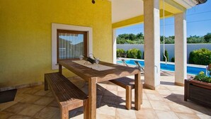 Een terras met een houten tafel en banken, een glazen deur en uitzicht op een zwembad.
